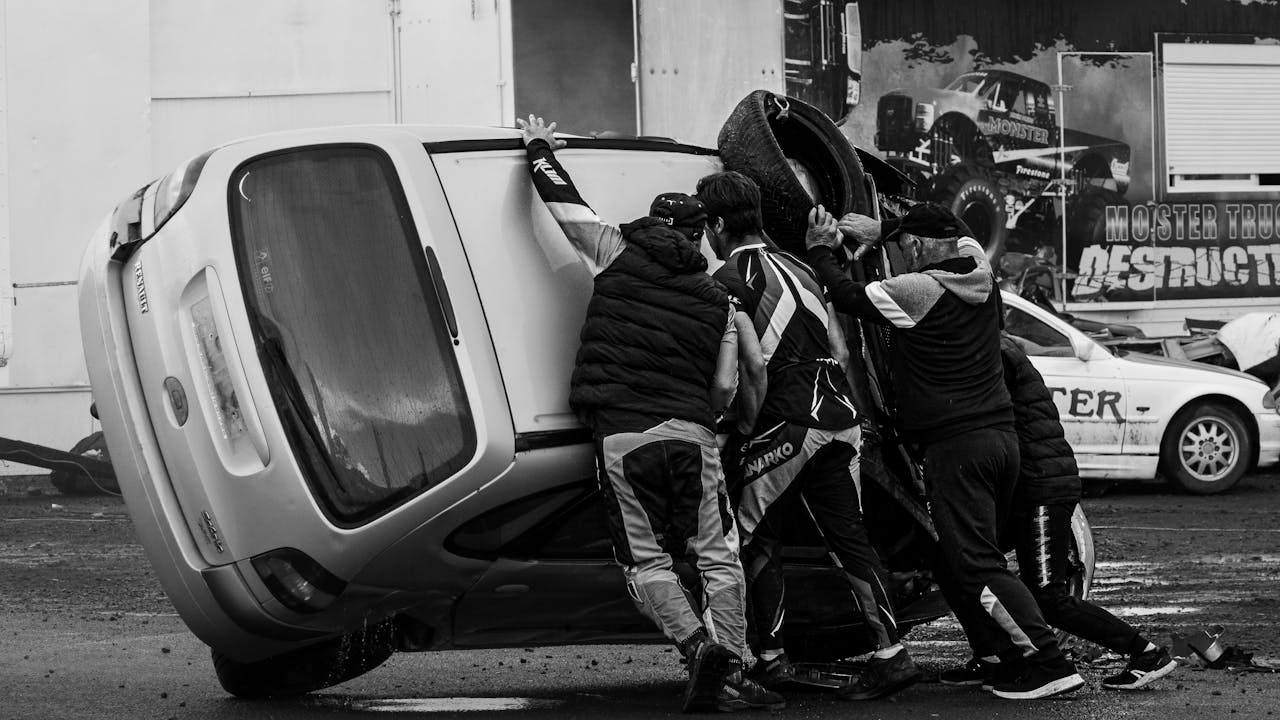 Black and white image of a team pushing an overturned car during a live stunt performance.